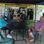 Groupe de personnes rassemblées sous un abri au golf des Quatre Saisons, discutant et se détendant autour d'une table. L'environnement est convivial, avec des chaises en plastique vert et des tables colorées. On aperçoit l'entrée du club et des voitures stationnées à l'extérieur, suggérant une ambiance de loisir et de camaraderie. Cette image illustre une journée typique au golf, où les passionnés se retrouvent pour partager des moments agré