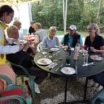 Groupe de personnes réunies autour d'une table à l'extérieur, profitant d'un repas convivial en pleine nature. Une jeune femme sert de la nourriture à un homme, tandis que d'autres convives discutent et dégustent divers plats. Des bouteilles de vin et des verres sont présents sur la table, indiquant une ambiance festive. L'environnement verdoyant et arboré crée une atmosphère agréable pour ce moment de partage et de convivialité.