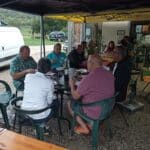 Groupe de personnes profitant d'un repas en plein air sous un abri, dans un cadre convivial. Ils partagent des plats et des boissons, créant une atmosphère chaleureuse. En arrière-plan, on aperçoit des véhicules et un paysage verdoyant, suggérant un lieu de détente à proximité de la nature. Cette image illustre des moments de convivialité et de partage, parfaits pour promouvoir des activités de restauration ou des événements en extérieur.