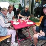 Groupe d'amis partageant un repas en plein air autour d'une table rouge. Deux femmes et deux hommes, souriants, sont assis sur des chaises en bois. Les plats et verres sur la table indiquent une ambiance conviviale. Un petit chien est également présent sous la table. L'environnement extérieur est verdoyant, suggérant un cadre agréable pour un repas en groupe.