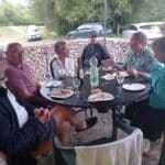 Groupe de personnes réunies autour d'une table en extérieur, partageant un repas convivial. On observe des plats et des boissons sur la table, ainsi qu'une ambiance détendue et amicale. En arrière-plan, des voitures sont garées sur un chemin bordé d'arbres, indiquant un cadre naturel propice aux rencontres. Cette image évoque des moments de partage et de convivialité en plein air, idéaux pour des événements sociaux ou des repas en groupe.