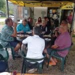Un groupe de personnes se retrouve autour d'une table en extérieur, profitant d'un repas convivial dans un cadre décontracté. Ils dégustent des plats variés et partagent des rires tout en sirotant des verres de vin. L'ambiance est animée, illustrant un moment de camaraderie entre amis ou membres d'un club, probablement après une activité sportive comme le golf. En arrière-plan, on aperçoit une voiture garée et des éléments de décoration liés au golf