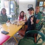 Quatre personnes profitent d'un repas convivial en extérieur, assises à une table colorée. L'ambiance décontractée est accentuée par des verres de vin et des plats savoureux. En arrière-plan, on aperçoit des éléments décoratifs et une enseigne du Golf des Quatre Saisons, indiquant que l'événement se déroule dans un cadre lié au golf. Les participants affichent des expressions joyeuses, suggérant un moment de partage et de camaraderie