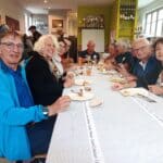 Un groupe de personnes réunies autour d'une table dans un cadre convivial, partageant un repas. Les convives, d'âges variés, affichent des sourires et discutent joyeusement. La table est garnie de plats, de boissons et de couverts, créant une ambiance chaleureuse et amicale. En arrière-plan, on aperçoit des trophées et des bouteilles, suggérant un lieu de rencontre pour des événements sociaux ou sportifs. Cette image illustre
