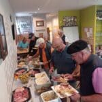 Un groupe d'hommes se regroupe autour d'un buffet dans un cadre convivial, où ils se servent divers plats. Sur la table, on aperçoit une variété de charcuteries, de salades, et d'accompagnements. L'ambiance est animée, avec des convives en discussion, créant une atmosphère chaleureuse. En arrière-plan, des trophées et des bouteilles sont visibles, témoignant d'un lieu de rencontre et de partage. Cette scène illustre un