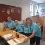 Quatre personnes souriantes, vêtues de polos turquoise, sont assises autour d'une table en bois dans un cadre convivial. Elles dégustent un repas composé de plats variés, entourées de décorations évoquant un club de golf. En arrière-plan, un tableau affiche les classements des championnats de golf. Cette image illustre une ambiance de camaraderie et de convivialité, typique des rencontres sportives.