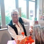 Employée souriante dans un café, ouvrant une bouteille tout en étant entourée de boissons et de matériel de service. Arrière-plan avec des fenêtres laissant passer la lumière naturelle et des affiches de menu. Lieu convivial, idéal pour se restaurer après une partie de golf. Ambiance chaleureuse et accueillante, mettant en avant le service et la satisfaction client.