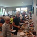 Groupe de personnes se servant à un buffet dans une salle conviviale. Des plats variés sont disposés sur une table, incluant des charcuteries et des accompagnements. En arrière-plan, d'autres convives sont assis et profitent de leur repas. L'ambiance est chaleureuse et animée, idéale pour des rencontres entre amis ou en famille.