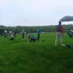 Un groupe de golfeurs s'entraîne sur un parcours de golf par temps nuageux. Certains joueurs utilisent des parapluies pour se protéger de la pluie légère. On peut voir des chariots de golf et des sacs de golf alignés sur le terrain. En arrière-plan, un bâtiment moderne servant de club-house est visible, entouré d'arbres verdoyants. Cette scène illustre une session d'entraînement de golf, mettant en avant la persévérance des