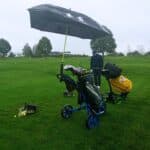 Un golfeur s'entraîne sur un terrain de golf par temps nuageux, utilisant un parapluie pour se protéger de la pluie. Deux chariots de golf sont visibles, l'un portant des clubs et des balles, et l'autre un équipement de marque TaylorMade. Des balles de golf sont éparpillées sur le sol, tandis que des arbres et des bâtiments se dessinent en arrière-plan, créant une ambiance typique de practice de golf.