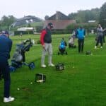 Groupe de golfeurs s'entraînant sur un terrain de golf sous un ciel nuageux. Les joueurs utilisent des chariots de golf pour transporter leurs clubs et se concentrent sur leur technique de swing. Des paniers de balles de golf sont disposés sur le gazon, tandis qu'un abri offre une protection contre la pluie. L'environnement verdoyant et les installations de golf en arrière-plan créent une atmosphère propice à l'apprentissage et à la pratique