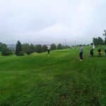 Un groupe de golfeurs se prépare à jouer sur un parcours verdoyant sous un ciel nuageux. L'un d'eux effectue un swing, tandis que d'autres attendent leur tour près de leurs chariots de golf. L'environnement calme et naturel, avec des arbres et des collines en arrière-plan, crée une ambiance propice à la pratique du golf. Cette image illustre l'esprit convivial et détendu du sport, même par temps couvert.