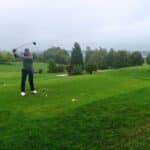 Un golfeur s'apprête à réaliser un coup sur un parcours verdoyant sous un ciel nuageux. Il est positionné sur le tee, prêt à frapper une balle de golf avec un driver. En arrière-plan, on aperçoit un drapeau sur un green, entouré d'arbres luxuriants et d'un paysage paisible. Cette image illustre parfaitement l'expérience relaxante et stratégique du golf en plein air.