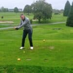Un golfeur se prépare à frapper sa balle sur un terrain de golf verdoyant. Habillé d'un pull gris et de pantalons foncés, il adopte une posture concentrée avant de réaliser son swing. En arrière-plan, on peut apercevoir des bunkers et des arbres, tandis que le temps est légèrement nuageux, ajoutant une ambiance calme à cette scène de golf. Cette image illustre parfaitement la passion du golf et l'importance de la technique dans
