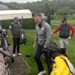 Trois golfeurs se préparent sur le parcours de golf par un temps nuageux. L'un d'eux, souriant, tient un club de golf tout en marchant. Les autres golfeurs, vêtus de vestes et de gilets, semblent discuter et se préparer pour leur partie. Les sacs de golf sont visibles à côté d'eux, témoignant de leur passion pour le sport. L'ambiance est conviviale et décontractée, typique des