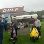 Groupe de golfeurs se préparant sur un parcours de golf sous un ciel nuageux et pluvieux. Des joueurs, munis de leurs chariots de golf, s'abritent sous des parapluies, tandis que le bâtiment en arrière-plan affiche le mot "GOLF". L'ambiance reflète la passion pour le sport malgré les conditions météorologiques difficiles, illustrant une journée typique sur le green.