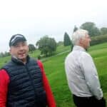 Deux golfeurs se tiennent sur un terrain de golf par temps nuageux. L'un porte une casquette et un gilet noir, affichant une expression amusée, tandis que l'autre, en arrière-plan, est vêtu d'une veste claire et regarde vers le terrain. L'environnement verdoyant et les éléments de golf visibles suggèrent une journée de jeu. Cette image illustre l'esprit convivial du golf et l'interaction entre les joueurs.