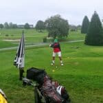 Un golfeur se prépare à frapper sa balle sur un parcours de golf verdoyant, sous un ciel nuageux. Il est vêtu d'une veste rouge et d'un pantalon blanc, portant également une casquette verte. À ses côtés, des sacs de golf sont visibles, témoignant de l'équipement nécessaire pour une partie. Le terrain présente des zones de sable et des greens, offrant un cadre idéal pour les amateurs de golf. Cette image illustre parfaitement l'ambiance d'une