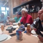 Groupe d'hommes autour d'une table dans un cadre convivial, partageant des boissons. Ils semblent discuter et profiter d'une ambiance détendue après une activité sportive. Des coupes et trophées sont visibles en arrière-plan, suggérant un lien avec le sport ou une compétition. Sur la table, on peut voir des verres de bière, des assiettes et des éléments de décoration. Cette image illustre un moment convivial entre amis ou collègues, idéal pour des événements de team building ou