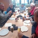 Groupe d'hommes réunis autour d'une table dans un cadre convivial, partageant des boissons et des plats. Certains portent des vêtements de sport, tandis que d'autres sont habillés de manière décontractée. La table est garnie de verres de bière, de plats vides et de serviettes. En arrière-plan, on aperçoit des trophées sur une étagère, suggérant une ambiance de compétition ou d'événements sportifs. L'image illustre un moment