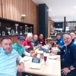 Groupe d'hommes réunis autour d'une table dans un cadre convivial, partageant des boissons, probablement après une partie de golf. En arrière-plan, des trophées brillants mettent en valeur des réalisations sportives. L'ambiance décontractée suggère une célébration ou un moment de camaraderie entre amis.