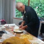 Homme en train de préparer des pommes de terre dans une cuisine lumineuse. Il coupe des rondelles de pommes de terre et les dispose dans un plat. Sur la table, on peut voir des ustensiles de cuisine, des ingrédients comme des oignons hachés, ainsi que des verres et un rouleau de papier essuie-tout. L'environnement est accueillant, avec des rideaux gris et une vue sur la verdure à l'extérieur. Cette scène illustre la préparation