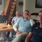 Quatre hommes se retrouvent autour d'une table en bois dans un intérieur chaleureux. Ils semblent engagés dans une conversation, chacun ayant une expression réfléchie. Des bouteilles de bière sont visibles sur la table, ajoutant une ambiance détendue. En arrière-plan, un escalier en bois mène à un étage supérieur, tandis qu'un tableau représentant un paysage est accroché au mur. Cette image capture un moment convivial entre amis, idéal pour illustrer des thèmes de sociabilité et de loisirs.