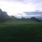 Image d'un parcours de golf au lever du soleil, montrant un green bien entretenu avec un drapeau blanc. En arrière-plan, des arbres verdoyants et un ciel nuageux créent une ambiance paisible. Cette scène évoque la tranquillité d'une matinée sur le parcours, idéale pour les amateurs de golf cherchant à profiter d'une partie matinale dans un cadre naturel.