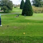Un golfeur se prépare à frapper sa balle depuis le tee sur un parcours de golf verdoyant. Il porte un polo bleu et un pantalon sombre, tenant un driver. En arrière-plan, d'autres golfeurs sont en action sur le green, tandis que des arbres et des collines verdoyantes entourent le terrain. Cette image illustre une journée agréable de golf, parfaite pour les amateurs de ce sport.