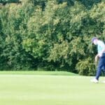 Golfeur en train de putter sur un green, avec le drapeau blanc et rouge en arrière-plan. La scène est entourée de verdure, illustrant un parcours de golf bien entretenu. L'image capture l'intensité et la concentration du joueur, mettant en avant l'aspect compétitif et stratégique du golf. Cette photographie évoque également une journée ensoleillée, idéale pour pratiquer ce sport.