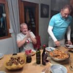 Repas convivial autour d'une table en bois, avec des plats de pâtes et de poulet frit. Deux hommes sont présents, l'un souriant et applaudissant, l'autre en train de servir. Plusieurs bouteilles de vin et de soda sont visibles, ainsi que des verres à vin. L'ambiance est chaleureuse et festive, idéale pour un moment entre amis ou en famille.
