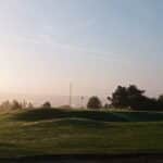 Paysage matinal d'un parcours de golf, avec une pelouse verte bien entretenue et des collines douces. En arrière-plan, des arbres et une antenne de communication se dessinent dans la brume, tandis qu'un drapeau de golf est visible sur le green. L'éclairage doux du matin crée une ambiance paisible, parfaite pour les amateurs de golf et les amoureux de la nature.