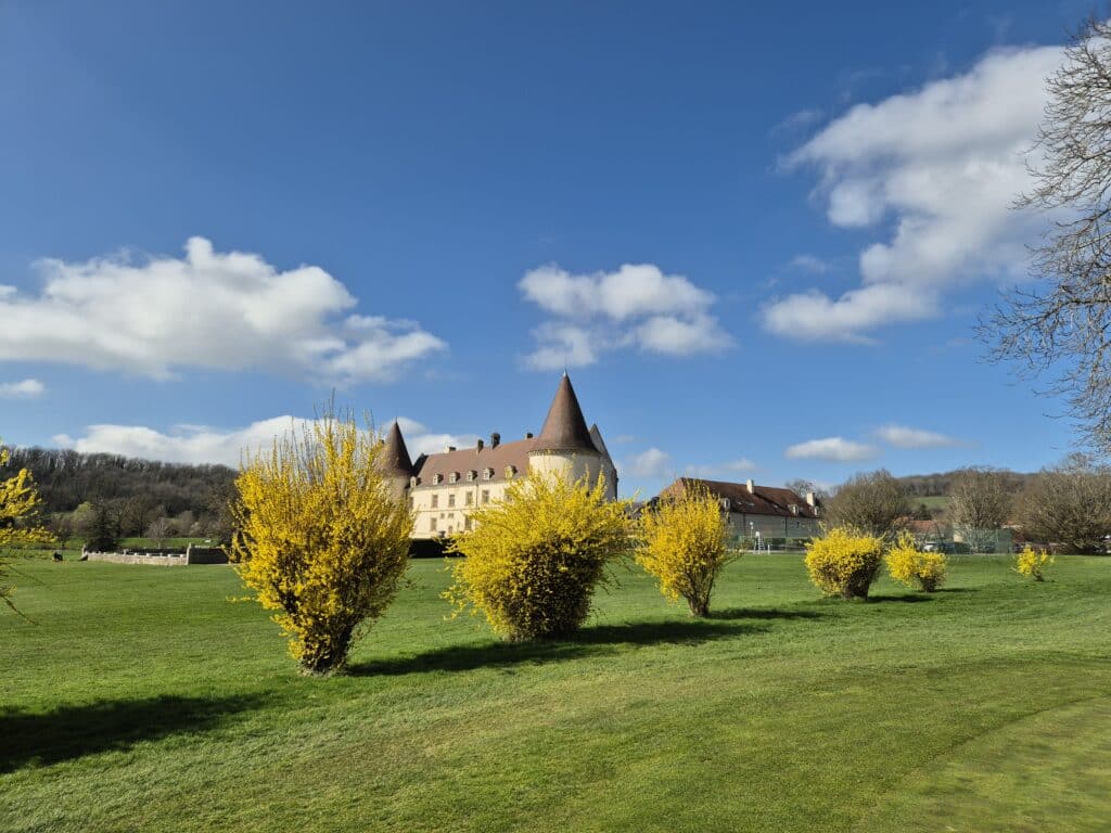 Château avec un jardin verdoyant et des arbustes jaunes en fleurs sous un ciel bleu. Le paysage inclut des collines en arrière-plan, créant une atmosphère paisible et pittoresque. Idéal pour les amateurs de nature et de photographie, ce site met en valeur l'architecture historique et la beauté des jardins printaniers.