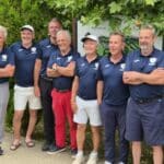 Groupe de seniors posant ensemble, vêtus de polos bleus portant le logo d'un club de sport. Ils se tiennent devant un panneau de bienvenue, entourés de verdure. L'image illustre l'esprit d'équipe et la camaraderie au sein de ce groupe dynamique, probablement lié à une activité sportive ou un événement communautaire. Cette photo met en avant la convivialité et l'engagement des membres.
