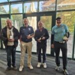 Quatre hommes souriants se tiennent debout à l'intérieur d'un bâtiment moderne avec de grandes fenêtres, portant des bouteilles de vin. L'un d'eux est vêtu d'un pull noir, tandis que les autres portent des vêtements de sport, dont un polo bleu. L'arrière-plan montre un espace lumineux avec des tables et des chaises, ainsi qu'un paysage verdoyant à l'extérieur. Cette image évoque une ambiance de convivialité et de célébration, probablement après
