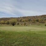 Vue panoramique d'un terrain de golf en automne, mettant en avant un green bien entretenu entouré de collines aux couleurs chatoyantes. Des golfeurs sont visibles en arrière-plan, profitant d'une journée ensoleillée. L'image illustre la beauté naturelle du paysage, avec des arbres aux feuilles dorées et une ambiance paisible, idéale pour les amateurs de golf et de nature.