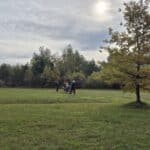 Groupe de personnes se rassemblant sur un terrain verdoyant, entouré d'arbres aux couleurs d'automne. Un ciel nuageux avec quelques éclaircies et un soleil partiellement visible. L'ambiance est conviviale, propice aux activités en plein air. Cette image évoque des moments de loisir et de détente en nature.