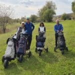 Groupe de quatre golfeurs sur un parcours verdoyant, équipés de chariots de golf. L'image montre un homme et trois femmes, souriants, prêts à jouer. En arrière-plan, on aperçoit des arbres aux feuilles automnales. Cette scène illustre une journée agréable de golf, mettant en avant l'esprit de camaraderie et l'amour du sport en plein air.