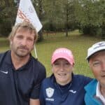 Groupe de trois golfeurs posant sur un parcours de golf, entourés d'arbres et de feuilles tombées au sol. L'un des golfeurs porte une casquette rose, tandis que les autres sont vêtus de polos et casquettes de sport. Le drapeau du parcours est visible en arrière-plan, indiquant une ambiance conviviale et sportive. Cette image évoque la passion du golf et les moments de partage entre amis sur le green.