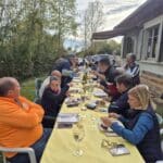 Repas convivial en plein air, avec un long tablée dressée sur une nappe jaune. Des convives dégustent des plats variés, autour de verres et de bouteilles. L'environnement naturel est agrémenté d'arbres en arrière-plan, et la maison au style rustique ajoute un charme unique à l'événement. Cette scène évoque des moments de partage et de convivialité entre amis ou en famille, idéale pour des rassemblements festifs.