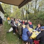 Groupe de personnes rassemblées autour d'une grande table en extérieur, profitant d'un repas convivial. L'événement se déroule près d'une maison, entourée d'une verdure automnale. Les participants, de différents âges, semblent apprécier la nourriture et les boissons, créant une ambiance chaleureuse et amicale. L'image illustre un moment de partage et de convivialité, idéal pour des activités de groupe ou des repas en plein air.