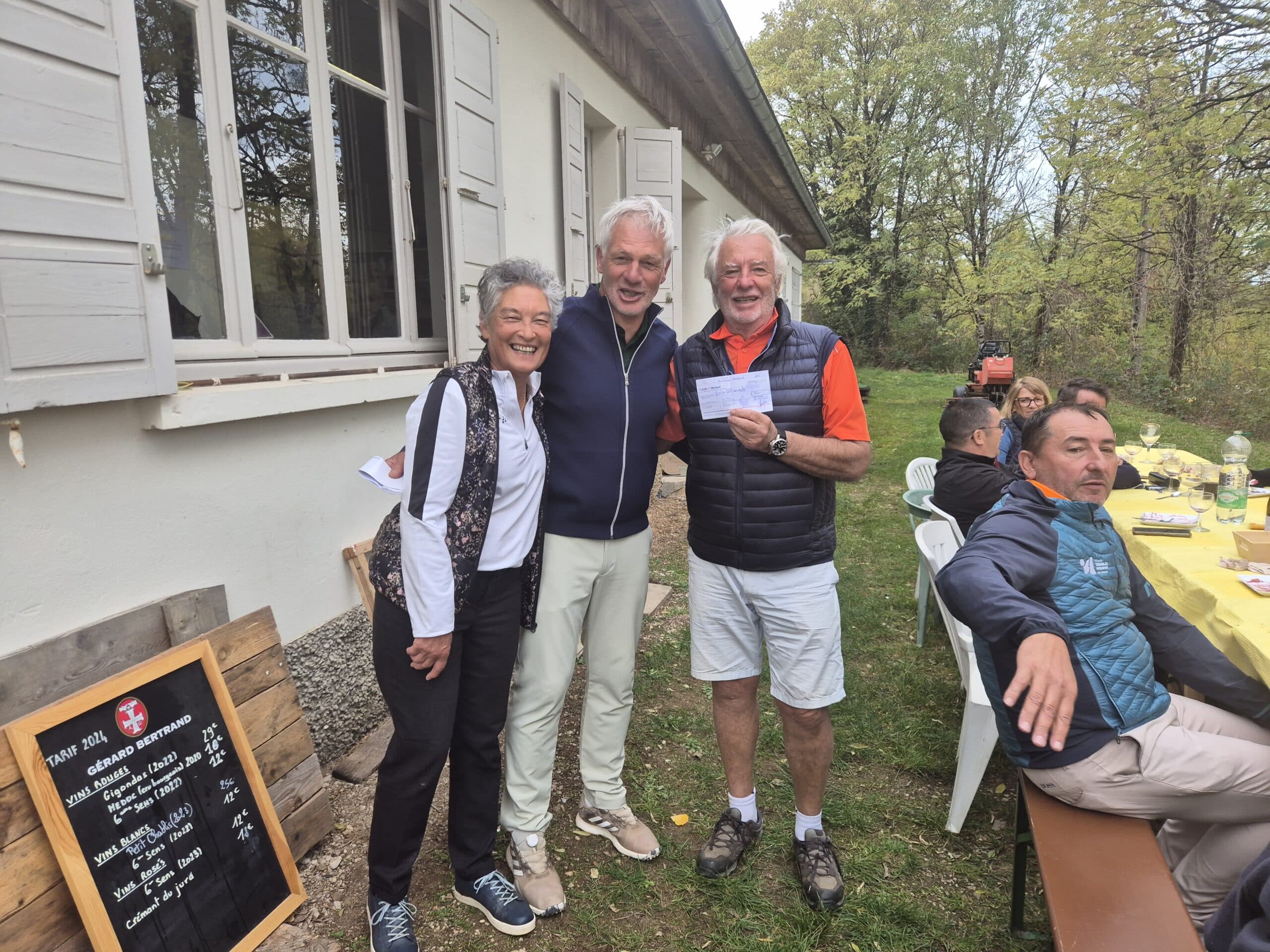 Groupe de personnes souriantes lors d'un événement en plein air, devant un bâtiment avec des fenêtres blanches. Trois individus posent ensemble, l'un d'eux tenant un chèque. En arrière-plan, une table est dressée avec des convives et des bouteilles, entourée d'arbres verdoyants. Un panneau affiche les tarifs des vins, ajoutant une touche conviviale à cette rencontre. L'image évoque une atmosphère de camaraderie et de gastronomie