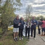 Quatre personnes posent ensemble dans un cadre naturel, entourées d'arbres et de verdure. Deux hommes et deux femmes sourient tout en tenant un prix ou un trophée. À l'arrière-plan, un stand est visible avec des assiettes et une nappe portant le logo d'une association, probablement liée à un événement communautaire ou sportif. L'atmosphère est conviviale et festive, suggérant une célébration ou une remise de prix.