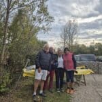 Quatre personnes se tiennent souriantes en plein air, entourées de verdure et d'un décor naturel. Elles sont réunies près d'une table recouverte d'une nappe jaune, où se trouvent des assiettes et des ustensiles. En arrière-plan, on aperçoit des voitures stationnées et un ciel nuageux, typique d'une journée d'automne. Cette image évoque un moment convivial en plein air, idéal pour des activités de groupe ou des rassemble