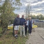 Groupe de quatre personnes souriantes posant en extérieur, entourées de verdure et de paysages naturels. Ils se tiennent près d'une table recouverte d'une nappe jaune, sur laquelle sont disposés des plats. L'arrière-plan présente des arbres feuillus et un ciel nuageux, créant une ambiance conviviale. Cette image évoque des moments de partage et de convivialité, idéale pour illustrer des activités en plein air ou des événements sociaux.
