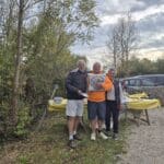Trois personnes posent ensemble lors d'un événement en plein air, entourées de verdure et de tables décorées de nappes jaunes. L'homme au centre porte un sweat orange et tient un objet, tandis que les deux autres sourient à ses côtés. L'arrière-plan montre des arbres aux feuilles d'automne et des véhicules stationnés. Cette image illustre un moment convivial, typique de rassemblements communautaires ou d'activités en plein air.