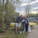 Trois personnes posent ensemble en plein air, entourées de verdure, lors d'un événement convivial. Elles sourient et semblent heureuses, tenant des objets dans leurs mains. À l'arrière-plan, une table recouverte d'une nappe jaune est disposée avec des ustensiles et des assiettes. Des véhicules sont garés à proximité, et le ciel est nuageux, ajoutant une atmosphère automnale à la scène. Cette image pourrait illustrer une rencontre