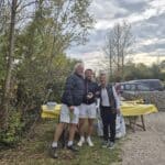 Trois personnes posent ensemble à l'extérieur, entourées d'une végétation automnale. L'une d'elles tient un prix ou un trophée, tandis qu'une table avec une nappe jaune est visible en arrière-plan, accompagnée de divers objets. Le paysage est calme, avec des voitures stationnées à proximité et un ciel nuageux. Cette image capture un moment convivial, probablement lors d'un événement ou d'une compétition en plein air.