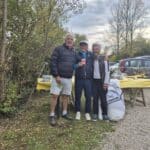 Photo de trois personnes souriantes posant ensemble en plein air, entourées de verdure et d'un décor convivial. L'un des individus porte un gilet noir et tient un petit drapeau canadien, tandis que les autres sont vêtus de vêtements décontractés. En arrière-plan, on remarque une table avec une nappe jaune et divers objets, ainsi qu'une voiture stationnée. Cette image évoque un moment de convivialité et de partage en milieu naturel.