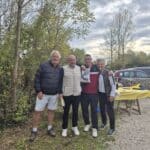 Un groupe de quatre personnes souriantes se tient ensemble en extérieur, entouré de verdure et d'arbres en arrière-plan. Ils portent des tenues décontractées adaptées à une journée en plein air, avec des vestes et des pantalons confortables. À l'arrière-plan, on aperçoit des voitures stationnées et une table avec une nappe jaune, suggérant une ambiance conviviale et festive. Cette image évoque des moments de camaraderie et de partage en pleine